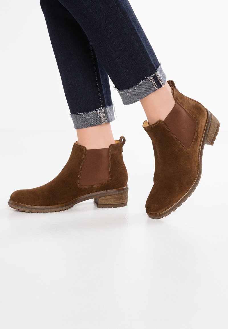 Pieds portant des bottines en suède marron avec des panneaux élastiques latéraux et des talons bas empilés, associées à un jean bleu foncé retroussé.