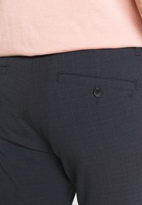 Marineblauwe broek van gestructureerde stof, met een enkele achterzak met een zwarte knoop en een subtiel gestreept patroon.