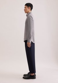 Camicia grigia chiaro con bottoni e maniche lunghe, abbinata a pantaloni blu navy a gamba dritta e scarpe nere stringate, vista di profilo.