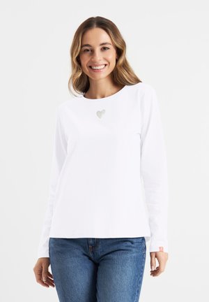 Witte langermige shirt met een klein hartontwerp op de borst, gecombineerd met blauwe jeans. Eenvoudige ronde halslijn en regular fit.