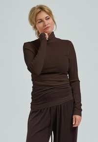 Kvinna med blont hår iklädd en brun polotröja och lösa byxor, som vilar hakan på sin hand medan hon tittar åt sidan.
