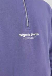 Fialová mikina s krátkým stříbrným zipem a bílým potiskem "Originals Studio" umístěným pod zipem na hrudi.