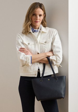 Frau trägt weiße Jeansjacke und blau gemustertes Halstuch, hält schwarze Tragetasche mit goldfarbenen Beschlägen und Logo.