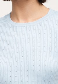 Κοντινό πλάνο ενός ατόμου που φορά ένα γαλάζιο ριμπ πλεκτό τοπ με μικρές λεπτομέρειες με οπές και στρογγυλή λαιμόκοψη με κυματιστό τελείωμα.