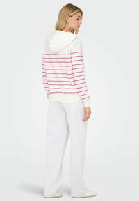 Felpa bianca con righe orizzontali rosa, dotata di cappuccio e polsini a costine. Abbbinata a pantaloni larghi grigi chiari e sneakers bianche.