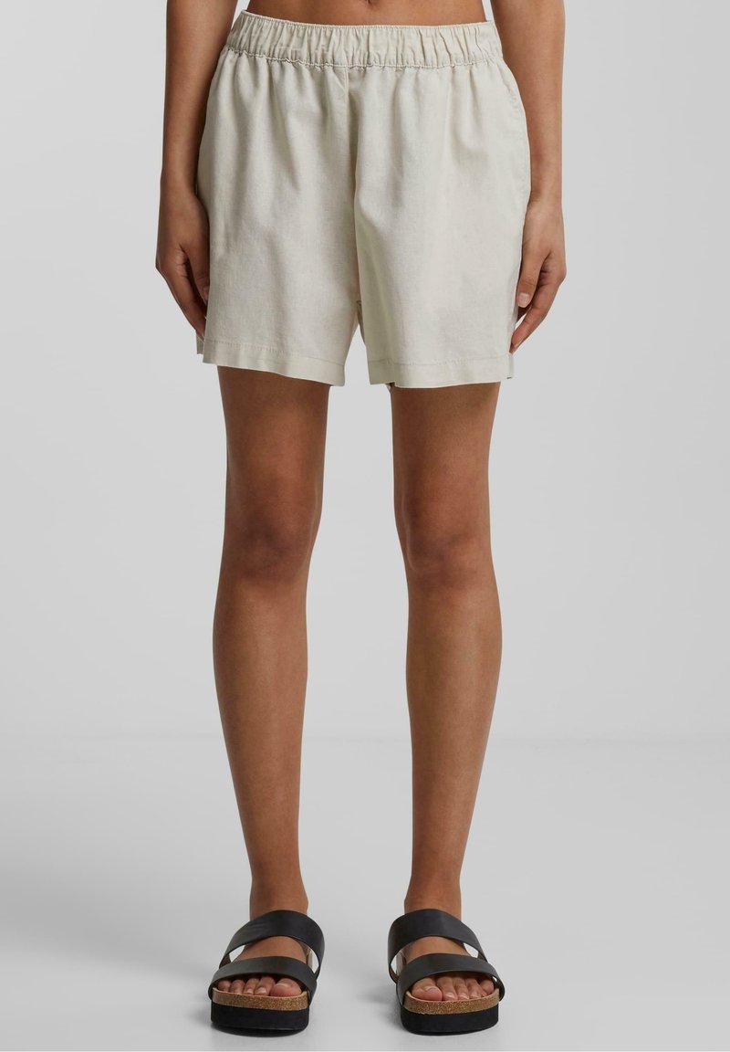 Personne debout portant un short beige à taille élastique et des sandales noires à double bride, montrée de la taille aux pieds sur un fond uni.