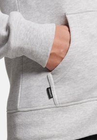 Sudadera gris claro con puños acanalados, un bolsillo frontal y una pequeña etiqueta negra en el lateral. Textura suave y diseño casual.