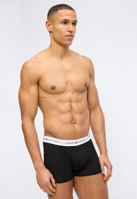 Άνδρας με μυώδες σώμα χωρίς πουκάμισο, που φοράει μαύρα μποξεράκια Tommy Hilfiger, στέκεται σε απλό ανοιχτό γκρι φόντο.