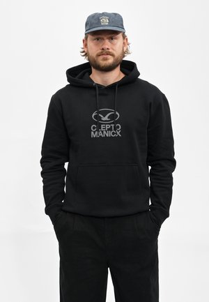 Mann mit Bart, der eine blaue Kappe und einen schwarzen Hoodie mit Fronttasche und Clepto Manicx-Logo trägt, steht mit den Händen in den Taschen.