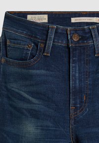 Jeans de mezclilla azul oscuro con cintura alta, que cuentan con costuras doradas, un pequeño bolsillo frontal y una cremallera con botón. La etiqueta dice "Levi's Premium."