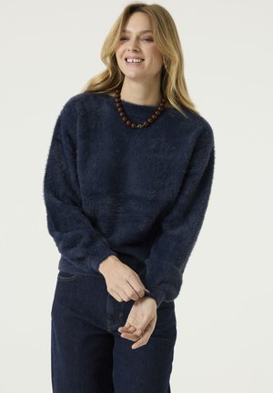 Young woman with long blonde hair wearing a navy blue fuzzy sweater and dark jeans, smiling and adjusting her sleeve against a plain background.