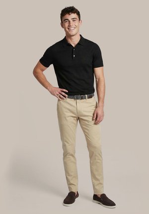Glimlachende man staand met hand in de zij, draagt een zwarte polo, beige broek, een riem met patroon en donkere instapschoenen tegen een beige achtergrond.