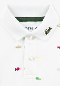 Polo blanc en tissu texturé, avec un col vert et des crocodiles brodés multicolores. Deux boutons au niveau de l'encolure.