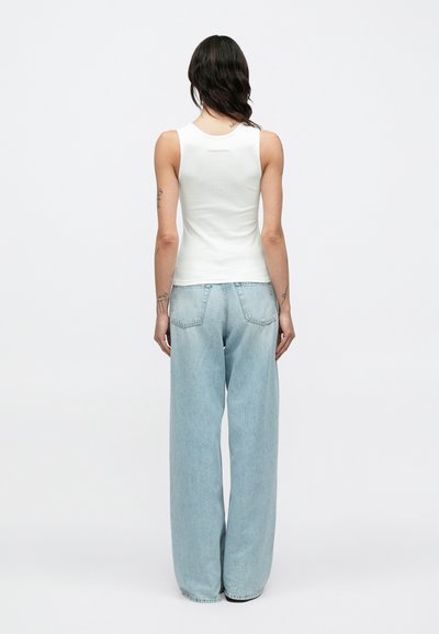 Λευκή εφαρμοστή φανέλα με στρογγυλή λαιμόκοψη, συνδυασμένη με ανοιχτόχρωμα μπλε τζιν παντελόνια με φαρδιά μπατζάκια. Το ύφασμα φαίνεται μαλακό με λεία υφή.