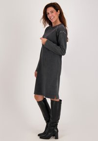 Vrouw die glimlacht, gekleed in een lange, donkergrijze jurk met studs op de schouders en zwarte laarzen met hoge hakken tot aan de knie, staande in profiel.
