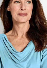 Blauw gedrapeerde top met een zachte textuur, met een wijde halslijn. Het model heeft lang, bruin haar en een gladde huid met zichtbare sproeten.