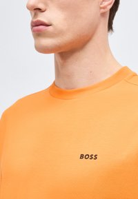 Κοντινό πλάνο ενός ατόμου που φοράει πορτοκαλί πουλόβερ με στρογγυλή λαιμόκοψη και τη λέξη "BOSS" τυπωμένη με μαύρα γράμματα στο στήθος.