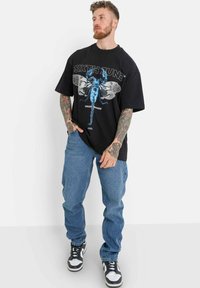 Svart t-shirt med blå grafik av en skorpion och texten "SIXTH TUNE." Matchas med ljusblå denimjeans och svartvita sneakers.