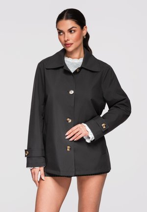 Femme aux cheveux foncés portant un manteau noir boutonné sur un pull blanc texturé, posant avec une main sur la taille devant un fond uni.