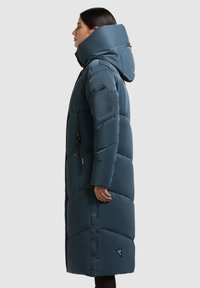 Lang, puffy pufferjas in donker teal met een hoge kraag en capuchon. Voorzien van ritstasjes en drukknoopsluiting, met een doorgestikt ontwerp.