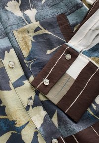 Chemise en textile à motif floral avec des couleurs bleu foncé, vert et marron. Elle présente des boutons contrastants et un tissu texturé.