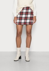 Jupe mini à carreaux rouge, vert et blanc, en tissu texturé ; associée à un pull en tricot blanc et des bottes beiges montantes.