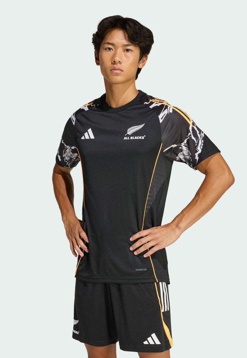 Černý sportovní dres s oranžovými akcenty a grafickým vzorem, v kombinaci s černými šortkami s bílými pruhy Adidas. Lehčí, prodyšný materiál.