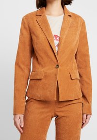 Kraftig orange kordflöjlsblazer med enkel knappstängning, slagna kragar, två framfickor och texturerad yta.