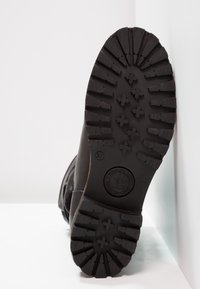 Semelle d'une botte noire montrant des crans profonds, des motifs en forme de croix, une inscription de taille 37 et un logo de marque circulaire près de la zone de la voûte plantaire.