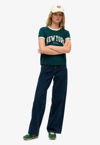 Camiseta verde con ribete blanco que presenta "NUEVA YORK" en impresión color crema. Pantalones vaqueros de pierna ancha en azul oscuro. Zapatillas verdes y rosas con una suela blanca.