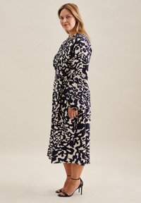 Robe longue à manches, de couleur marine avec un motif abstrait crème, cintrée à la taille. Portée avec des talons noirs à lanières, mettant en avant une texture lisse.