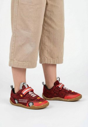 Kind draagt rode sneakers met brandweerwagen-thema en beige cropped broek op witte achtergrond.