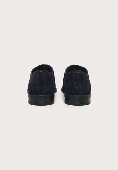 Paire de chaussures habillées en daim bleu marine vues de dos avec talons empilés noirs sur fond blanc.