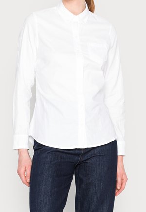 Femme portant une chemise blanche cintrée boutonnée avec un col et des manches longues, associée à un jean bleu foncé.
