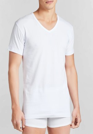 T-shirt bianca a scollo a V realizzata in morbido cotone, con maniche corte, taglio dritto e texture liscia, indossata sopra pantaloni corti bianchi.
