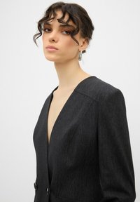 Donkergrijze blazer met een diepe V-hals, getailleerde pasvorm, scherpe schouders en twee grote knopen aan de voorkant voor sluiting.