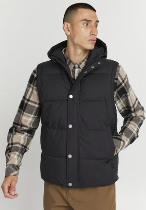 Svart puffervest med avtagbar hette, trykknapper og to sidelommer, iført over en rutete flanellskjorte med beige og svarte toner.