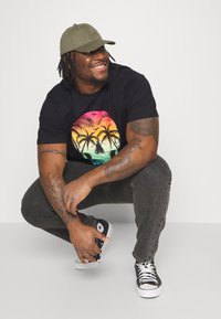 Black t-shirt featuring a colorful palm tree graphic, gray jeans, and black Converse sneakers. Olive green baseball cap worn slightly askew.