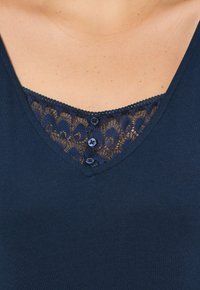 Námořnická modrá top s výstřihem do V, který obsahuje krajkovou vsadku se zvlněným okrajem a dvěma malými knoflíky na výstřihu. Hladká textura látky.