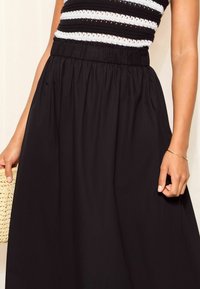 Černá dlouhá sukně s elastickým pasem, hladkou texturou a širokým střihem. Ruka drží tkanou psaníčko.