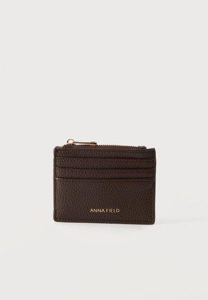 Porte-cartes en cuir texturé marron foncé avec trois emplacements pour cartes, un compartiment zippé et un logo doré "ANNA FIELD" à l'avant.