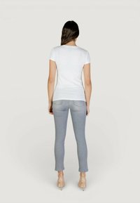 Vrouw met lang donker haar, staand met de rug naar de kijker, gekleed in een wit T-shirt met korte mouwen, lichtgrijze spijkerbroek en nude hoge hakken, tegen een effen achtergrond.