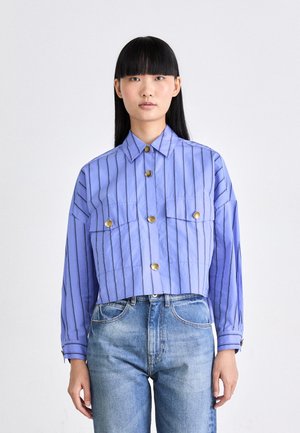 Camicia a maniche lunghe di colore viola chiaro con strisce verticali, due tasche frontali e bottoni dorati; abbinata a jeans in denim blu.