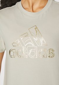 Lys grå T-shirt lavet af bomuld med en rund halsudskæring. Har et stort metalisk guld- og hvidt Adidas-logo med geometriske mønstre.