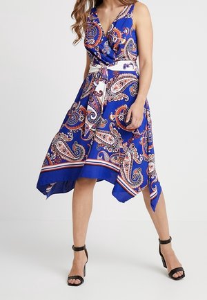Niebieska sukienka w stylu paisley z kopertowym fasonem, bez rękawów i asymetrycznym dołem. Z białym paskiem i misternymi wzorami w kolorze czerwonym i kremowym.