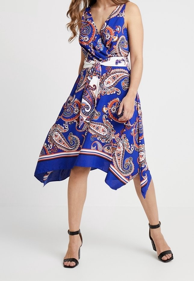 Robe bleu à motif cachemire avec un design portefeuille, sans manches et un ourlet asymétrique. Elle comporte une ceinture blanche et des motifs complexes en rouge et crème.