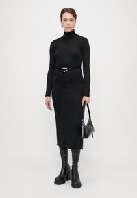 Schwarzes Langarmkleid mit hohem Kragen und einem Gürtel mit goldener Schnalle. Kombiniert mit knielangen schwarzen Stiefeln und einer kleinen schwarzen Handtasche.