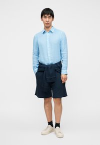 Γαλάζιο λινό πουκάμισο με κουμπιά, σκούρα μπλε σορτς με δέσιμο στη μέση, εκρού αθλητικά παπούτσια και μαύρες κάλτσες. Μινιμαλιστική, καθημερινή εμφάνιση.