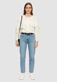 Blusa bianca con volant e maniche lunghe, abbinata a jeans chiari a vita alta e sneakers bianche. Inclusi cintura nera e borsa a spalla.