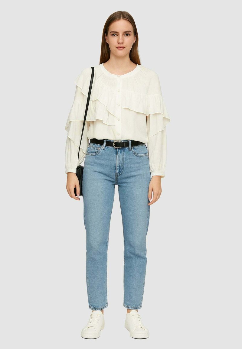 Blusa bianca con volant e maniche lunghe, abbinata a jeans chiari a vita alta e sneakers bianche. Inclusi cintura nera e borsa a spalla.